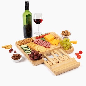 May include: A bamboo cheese board with a built-in drawer for cheese knives. The board has compartments for crackers, grapes, cheese, and salami. There is a bottle of wine, a glass of red wine, and a bowl of almonds in the image.