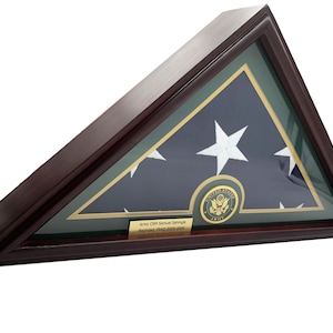 Könnte beinhalten: Eine hölzerne Schattenbox mit einer gefalteten amerikanischen Flagge im Inneren. Die Flagge wird mit einem grünen und goldenen Rand präsentiert. Eine goldene Plakette mit dem Text "Army CSM Samuel Schryver Baghdad, Iraq 2003-2005" ist an der Unterseite der Box angebracht. Ein goldenes Siegel mit den Worten "United States Army" ist auf der Flagge sichtbar.