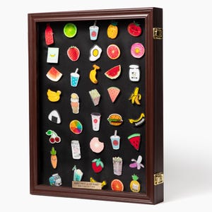 May include: A dark wood display case with a black interior containing a collection of colorful enamel pins shaped like various foods, fruits, and drinks. A small gold plaque at the bottom reads RARE FRUIT & GOURMET PIN COLLECTION.