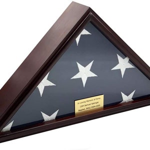 Puede incluir: Una caja de sombra triangular de madera con una bandera estadounidense doblada en su interior. La caja tiene una placa dorada con la inscripción "In Loving Memory of Army CSM Samuel Schringele Baghdad, IRAQ 2004-2005".
