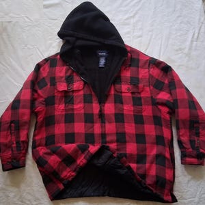 Puede incluir: Chaqueta con capucha a cuadros rojos y negros con interior forrado de forro polar negro. La chaqueta tiene cierre de cremallera completa, dos bolsillos en el pecho y un dobladillo inferior acolchado. La capucha es negra.