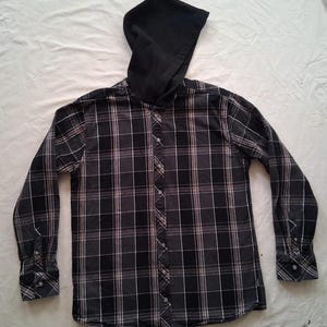 Puede incluir: Una camisa a cuadros negros y grises con capucha negra. La camisa presenta un patrón de cuadros clásico en tonos de negro, gris y blanco. Las mangas largas tienen puños abotonados. La capucha es de color negro liso.