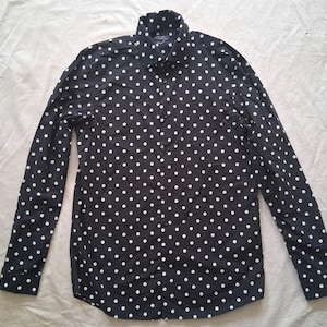 Puede incluir: Camisa de manga larga azul marino con un estampado de lunares blancos. La camisa tiene un cuello clásico y puños abotonados. La tela parece ser un material ligero, adecuado para diversas ocasiones.