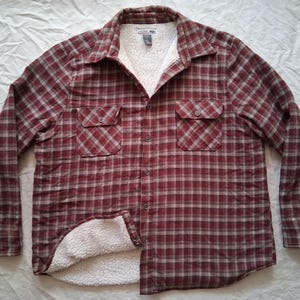 May include: A red and gray plaid flannel shirt with a white sherpa lining. The shirt has two chest pockets and a button-down front. The long-sleeved shirt is made of a soft, warm material.