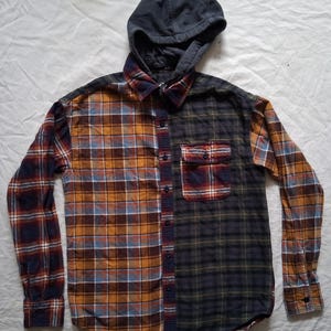 May include: A button-down flannel shirt with a navy blue hood. The shirt is split vertically, with a plaid pattern in shades of orange, blue, and navy on one side, and a darker green and navy plaid on the other. A pocket is on the left side.