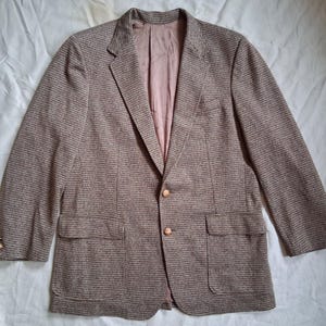 May include: A brown blazer with a textured pattern. The blazer has two front pockets, a notched lapel, and two gold-colored buttons. The interior lining is a light pink color.