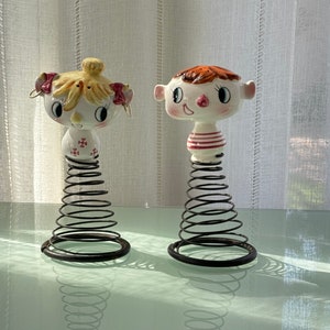 Könnte beinhalten: Zwei Keramikfiguren eines Mädchens und eines Jungen mit bemalten Gesichtern und Haaren. Sie stehen auf federbelasteten Sockeln und sind weiß mit roten und rosa Akzenten.