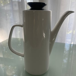 Könnte beinhalten: Weiße Kaffeekanne aus Keramik mit schwarzem Deckel. Die Kanne hat eine hohe, zylindrische Form und einen Ausguss an der Seite. Der Griff ist an der Seite der Kanne befestigt.