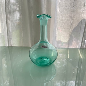 Op de afbeelding: Een lichtgroene glazen karaf met een gekrulde bovenkant en een label met de tekst "Bleuvert Handcraft".