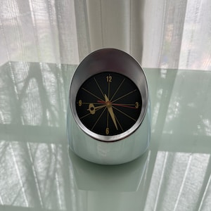 May include: A silver and black analog clock with a round face and gold accents. The clock has a unique design with a curved base and a raised face.