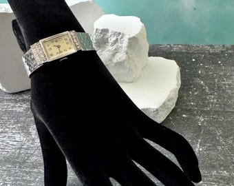 May include: A silver-toned wristwatch with a rectangular face and a band adorned with small, sparkling embellishments. The watch is displayed on a black velvet hand-shaped display against a backdrop of white stone and a teal-colored wall.