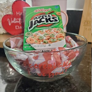 Peut inclure: Un bol en verre transparent avec le texte "I cereal-sly love you" en lettres rouges, rempli de bonbons emballés. Une boîte de céréales Kellogg's Apple Jacks est à l'intérieur du bol. Une décoration en forme de cœur rouge est en arrière-plan.