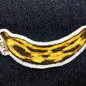 May include: A yellow and brown embroidered banana patch with the word "DYE" stitched on the side.