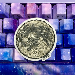 May include: Embroidered patch featuring a detailed, grayscale depiction of the moon with a textured surface and a white border. The patch is set against a keyboard with purple and blue keys. The text "OYE" is visible.