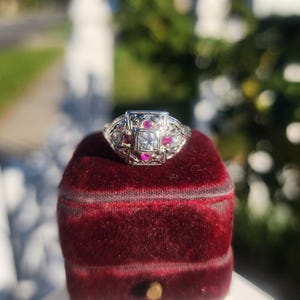 May include: A vintage-style ring with a square, central diamond, surrounded by smaller diamonds and vibrant red gemstones. The ring is set in a detailed, ornate metalwork design, resting on a deep red velvet ring box.