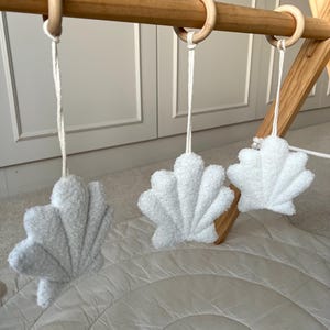 K&ouml;nnte beinhalten: Drei wei&szlig;e, muschelf&ouml;rmige H&auml;ngeornamente aus einem strukturierten Pl&uuml;schstoff. Jede Muschel ist an einer wei&szlig;en Kordel an einem glatten, hellen Holzring befestigt, der an einer horizontalen Holzstange eines Spielbogens angebracht ist.