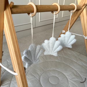K&ouml;nnte beinhalten: Ein h&ouml;lzernes Spieltrapez f&uuml;r Babys mit drei hellen, strukturierten, muschelf&ouml;rmigen H&auml;ngespielzeugen, die an glatten Holzringen befestigt sind. Das Gestell besteht aus hellem Holz und wird durch eine weiche, runde, hellgraue Spielmatte erg&auml;nzt.