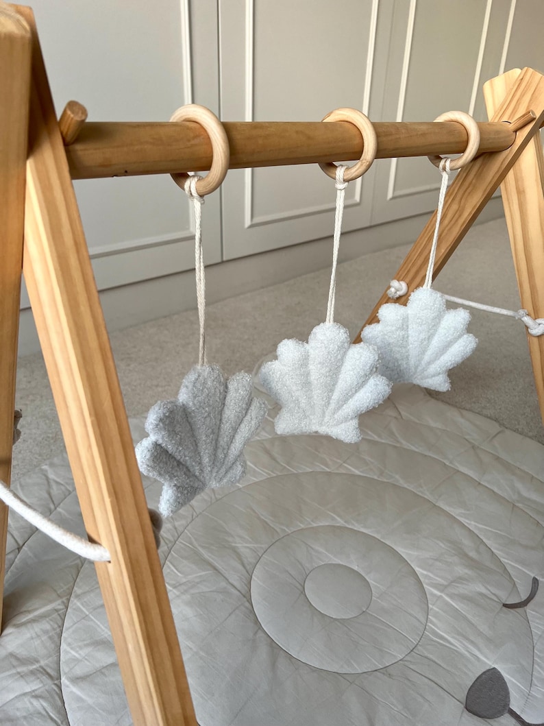 K&ouml;nnte beinhalten: Ein h&ouml;lzernes Spieltrapez f&uuml;r Babys mit drei hellen, strukturierten, muschelf&ouml;rmigen H&auml;ngespielzeugen, die an glatten Holzringen befestigt sind. Das Gestell besteht aus hellem Holz und wird durch eine weiche, runde, hellgraue Spielmatte erg&auml;nzt.
