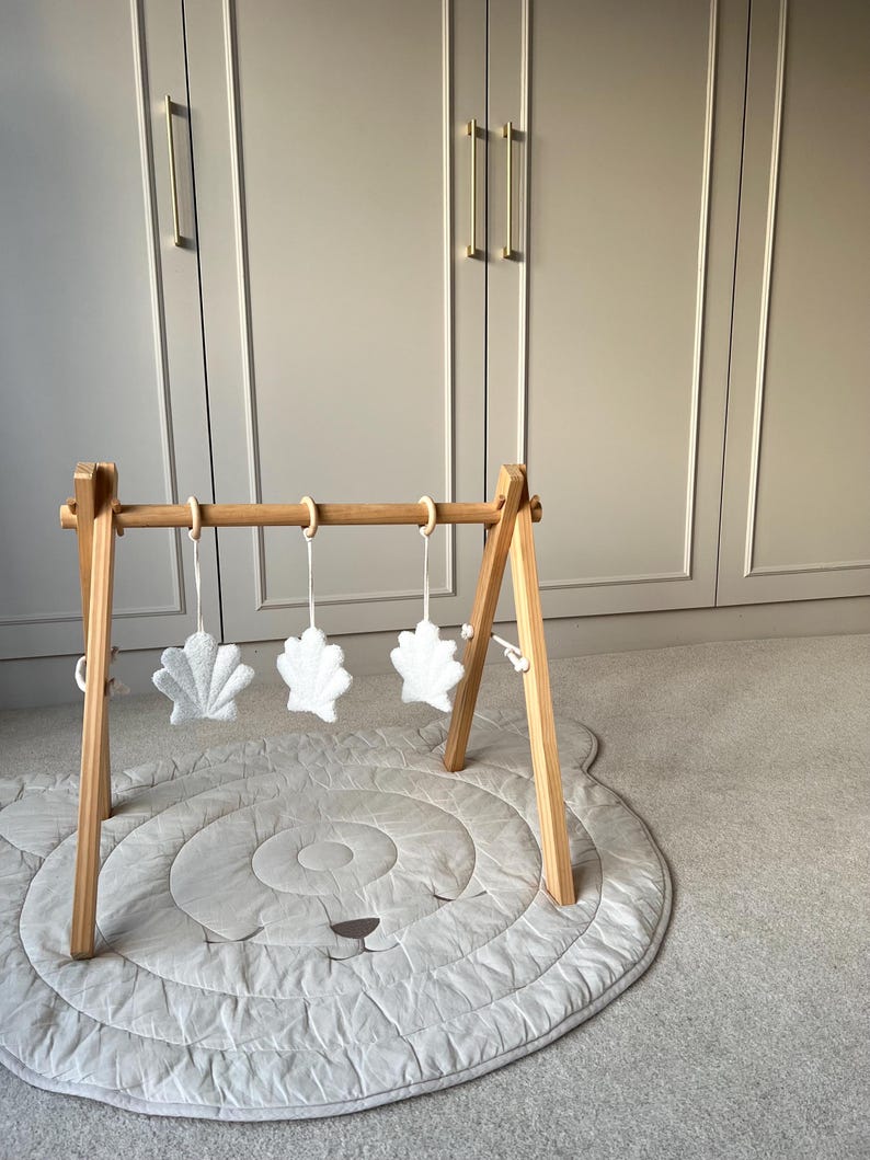 K&ouml;nnte beinhalten: Ein h&ouml;lzernes Spieltrapez mit einem hellen Rahmen und drei h&auml;ngenden, wei&szlig;en, muschelf&ouml;rmigen Pl&uuml;schspielzeugen, die an Holzringen befestigt sind. Das Spieltrapez steht auf einer runden, gesteppten, hellgrauen Spielmatte mit aufgesticktem Gesicht.