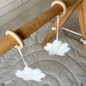 K&ouml;nnte beinhalten: Ein h&ouml;lzernes Spieltrapez f&uuml;r Babys mit einer horizontalen Stange und zwei glatten Holzringen. An diesen Ringen h&auml;ngen zwei wei&szlig;e, strukturierte, blattf&ouml;rmige Stoffanh&auml;nger an wei&szlig;en Schn&uuml;ren &uuml;ber einer gesteppten, kreisf&ouml;rmigen Spielmatte.