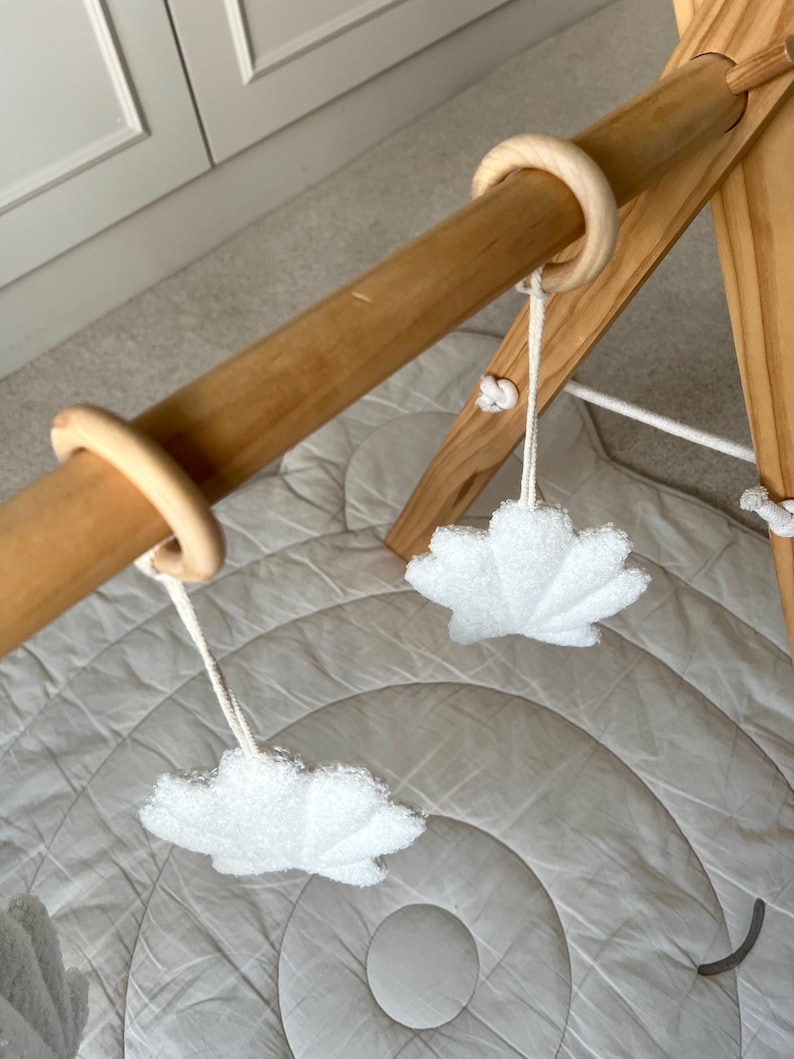 K&ouml;nnte beinhalten: Ein h&ouml;lzernes Spieltrapez f&uuml;r Babys mit einer horizontalen Stange und zwei glatten Holzringen. An diesen Ringen h&auml;ngen zwei wei&szlig;e, strukturierte, blattf&ouml;rmige Stoffanh&auml;nger an wei&szlig;en Schn&uuml;ren &uuml;ber einer gesteppten, kreisf&ouml;rmigen Spielmatte.
