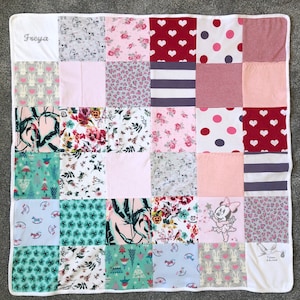 Op de afbeelding: Een patchwork quilt met een witte rand. De quilt bestaat uit vierkanten met verschillende patronen, waaronder bloemen, harten, stippen en dierenprints in roze, rood, grijs en wit. De naam "Freya" is in grijs op een witte hoek geborduurd.