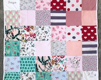 May include: A patchwork quilt with a white border. The quilt is made up of squares with various patterns, including floral, hearts, polka dots, and animal prints in pink, red, grey, and white. The name "Freya" is embroidered in grey on a white corner.