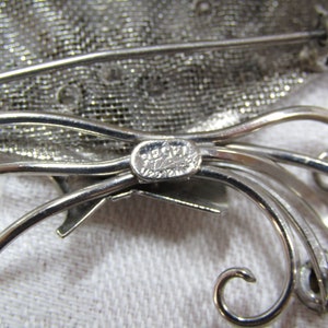 May include: Silver tone brooch with a mesh design and a floral motif. The brooch is marked "Piccadilly 1/20/12K".