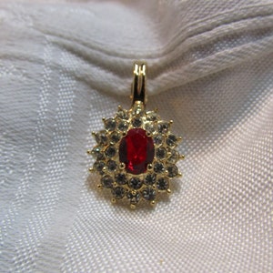 Pode incluir: Um pingente de ouro com uma pedra oval vermelha cercada por strass transparentes.