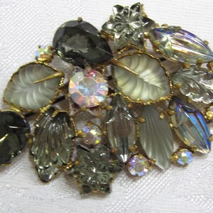 Könnte beinhalten: Eine Vintage-Brosche mit einem Cluster aus irisierenden und undurchsichtigen Glassteinen in Grün-, Grau- und Weißtönen. Die Steine sind in goldfarbenem Metall gefasst und haben verschiedene Formen, darunter Blätter, Blumen und geometrische Formen.