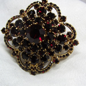 Peut inclure: Une broche dorée ornée d'un groupe de pierres circulaires facettées rouge foncé disposées selon un motif symétrique et complexe. La monture en métal est travaillée en filigrane et maintient fermement de nombreuses gemmes rouges.