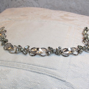 May include: A silver-toned bracelet with clear, sparkling rhinestones and decorative metalwork. The bracelet features a series of oval and leaf-shaped elements, connected by delicate links. The rhinestones are set in clusters and individual settings.