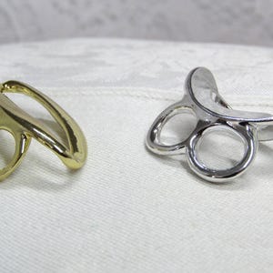 May include: Two metal rings, one gold-toned and one silver-toned, each featuring a smooth, curved, open-frame design with two circular openings at the base and a wider, arched top section that extends outward, creating a distinct, sculptural shape.