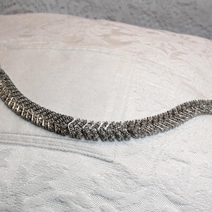 Op de afbeelding: Een zilverkleurige armband met een chevronontwerp. De armband bestaat uit meerdere rijen kleine, rechthoekige schakels, waardoor een textuur ontstaat. De sluiting is aan één uiteinde zichtbaar. Op een witte, getextureerde ondergrond.