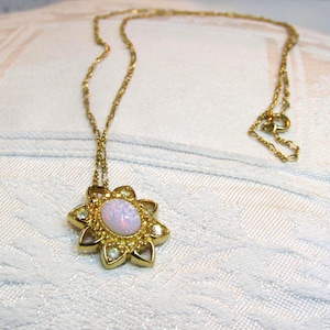 May include: A gold-toned necklace featuring a flower-shaped pendant with a central oval opal stone. The pendant is surrounded by smaller, clear stones. The necklace chain is delicate and has a clasp closure.