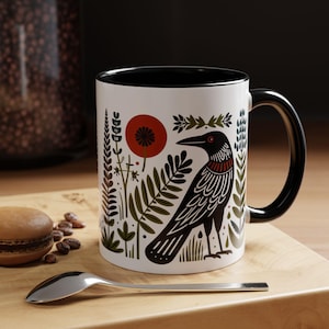 May include: White ceramic mug with a black interior and handle. The mug features a black raven illustration with red accents, surrounded by a floral design in green and red. A spoon and a pastry are in the foreground.