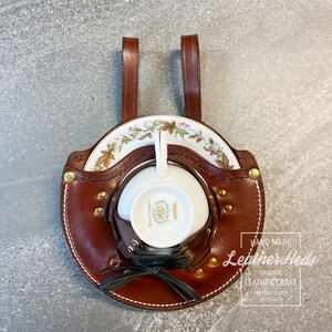 Pu&ograve; includere: Un porta tazza e piattino in pelle marrone con due cinturini. Il porta tazza ha un design con lacci e rivetti dorati. Il porta tazza &egrave; etichettato "Hand Made Leather Heds Original Leather Craft Mooree Beach Australia".