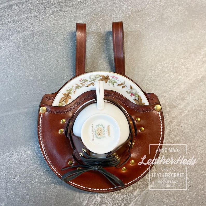 Pu&ograve; includere: Un porta tazza e piattino in pelle marrone con due cinturini. Il porta tazza ha un design con lacci e rivetti dorati. Il porta tazza &egrave; etichettato "Hand Made Leather Heds Original Leather Craft Mooree Beach Australia".