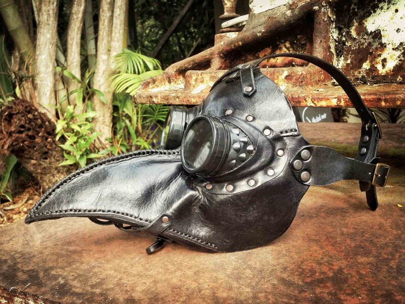 May include: A black leather plague doctor mask with a round, clear eye piece and a long, curved beak. The mask is decorated with metal studs and rivets.