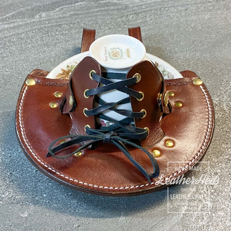 Pu&ograve; includere: Un porta tazze in pelle marrone con una tazza da t&egrave; e un piattino bianchi all'interno. Il porta tazze ha un design con lacci con lacci neri e accenti in metallo dorato. Il testo "HAND MADE Leathercraft ORIGINAL LEATHER CRAFT" &egrave; visibile nell'angolo in basso a destra dell'immagine.