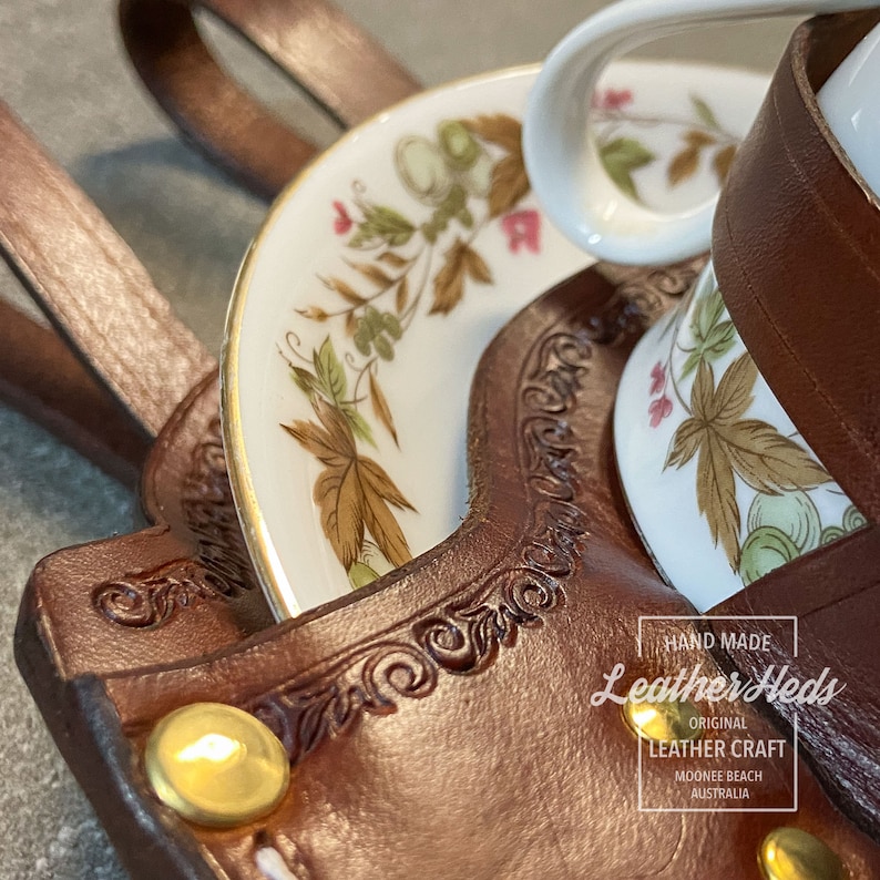 Pu&ograve; includere: Un porta tazza e piattino in pelle marrone con un bottone dorato e il testo "Leather Heds Original Leather Craft Moonie Beach Australia".