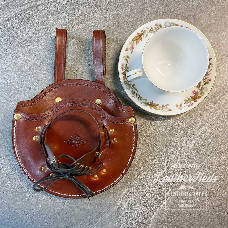 Pu&ograve; includere: Un porta tazze in pelle marrone con due anelli di sospensione e un design decorativo. Il porta tazze &egrave; realizzato da Leatherfields Original Leather Craft, Mooloolaba, Australia.
