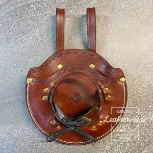 Pu&ograve; includere: Un fodero in pelle marrone con un design decorativo e dettagli in ottone. Il fodero ha due cinghie per appendere e una chiusura con lacci. Il testo "Handmade Leathercraft" &egrave; visibile nell'angolo in basso a destra.