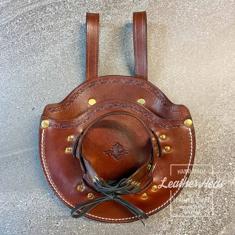 Pu&ograve; includere: Un fodero in pelle marrone con un design decorativo e dettagli in ottone. Il fodero ha due cinghie per appendere e una chiusura con lacci. Il testo "Handmade Leathercraft" &egrave; visibile nell'angolo in basso a destra.