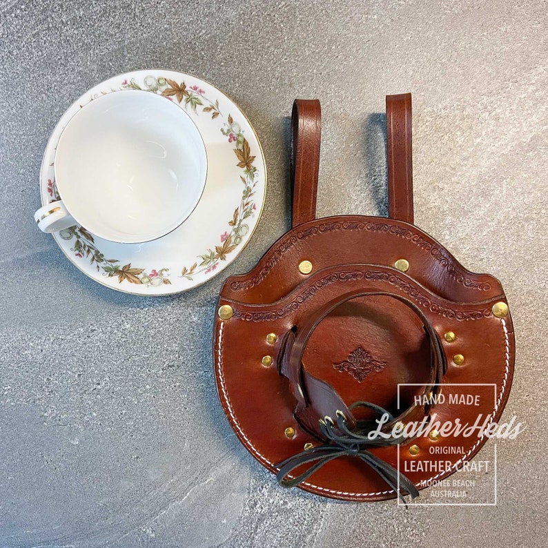 Pu&ograve; includere: Un porta tazze in pelle marrone con un bordo bianco cucito e rivetti in ottone. Il porta tazze ha due cinturini in pelle per appenderlo. Il testo "HAND MADE Leather Heads ORIGINAL LEATHER-CRAFT MOOMEE BEACH AUSTRALIA" &egrave; stampato su un'etichetta bianca.