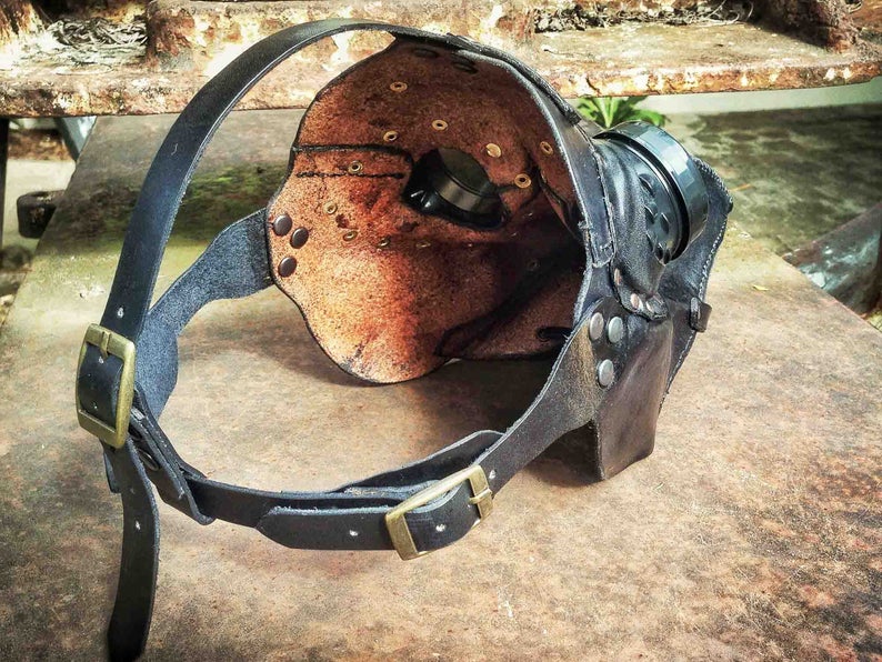 May include: A black leather steampunk-style mask with a brass buckle and rivets. The mask has a round, open eye area and a small, round lens on the side.