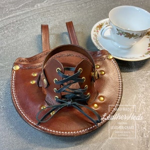 Pu&ograve; includere: Un porta tazze in pelle marrone con chiusura a lacci neri e dettagli in ottone. Il porta tazze ha due cinghie per il trasporto. Il testo "Hand Made Leather Heds Original Leather Craft Mooloolaba Beach Australia" &egrave; stampato sul porta tazze.