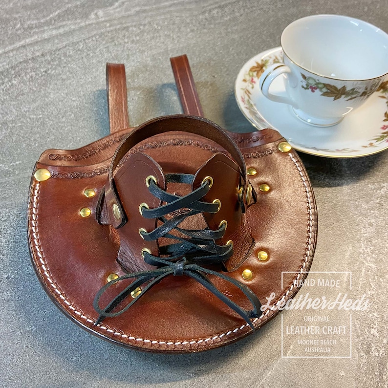 Pu&ograve; includere: Un porta tazze in pelle marrone con chiusura a lacci neri e dettagli in ottone. Il porta tazze ha due cinghie per il trasporto. Il testo "Hand Made Leather Heds Original Leather Craft Mooloolaba Beach Australia" &egrave; stampato sul porta tazze.