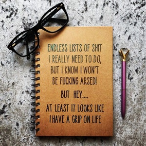 May include: A spiral-bound notebook with the text "ENDLESS LISTS OF SHIT I REALLY NEED TO DO, BUT I KNOW I WON'T BE FUCKING ARSED! BUT HEY.... AT LEAST IT LOOKS LIKE I HAVE A GRIP ON LIFE." Black eyeglasses and a purple pen with a diamond-shaped top are also in the image.