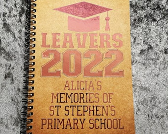 Pode incluir: Um caderno espiral marrom com um chapéu de formatura rosa e o texto "Leavers 2022" em vermelho. O texto "Alicia's Memories of St Stephen's Primary School" também está em vermelho.